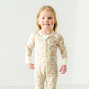 A young toddler with blonde hair smiling at the camera, wearing a Makemake Organics Organic 2-Way Zip Romper - Summer Floral with a white collar, standing against a plain white background.