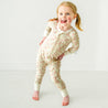 A young, happy toddler in a Makemake Organics Organic 2-Way Zip Romper - Summer Floral playfully poses with one hand on her hip in a brightly lit room with a white background.