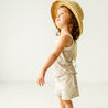 A joyful toddler girl wearing a Makemake Organics Organic Spaghetti Top & Shorts Set - Summer Floral and a straw hat smiling and twirling in a bright, white room.