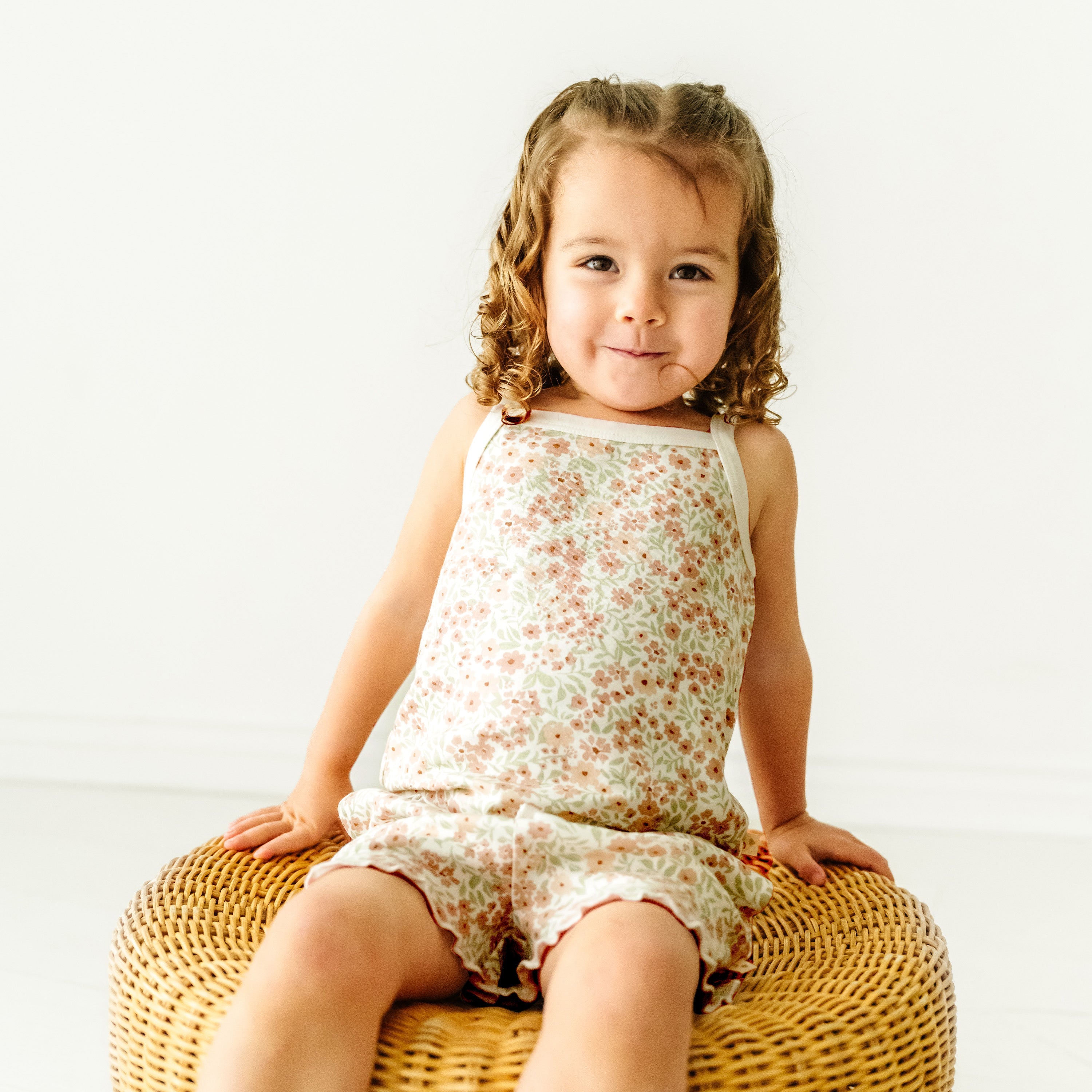 A young child with curly hair sits on a woven stool, wearing a Makemake Organics Organic Spaghetti Top & Shorts Set - Summer Floral, smiling playfully at the camera in a bright, white room.