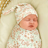 A newborn baby sleeps swaddled in a Makemake Organics Organic Swaddle Blanket & Hat - Summer Floral outfit and matching hat, resting in a woven basket with a soft yellow background.
