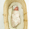 A newborn baby sleeps peacefully in a wicker bassinet, swaddled in a Makemake Organics Organic Swaddle Blanket & Hat - Summer Floral matching their hat. the bassinet is lined with a soft, cream-colored pad.