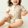 A cheerful toddler girl in a Makemake Organics Organic Short Zip Romper - Summer Floral sits on a light wooden floor, laughing with her mouth open in a bright, airy room.