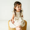 A young toddler with dark hair, wearing a Makemake Organics Organic Flutter Dress in Summer Floral, sits on a wicker stool in a bright room, looking to the side with a thoughtful expression.