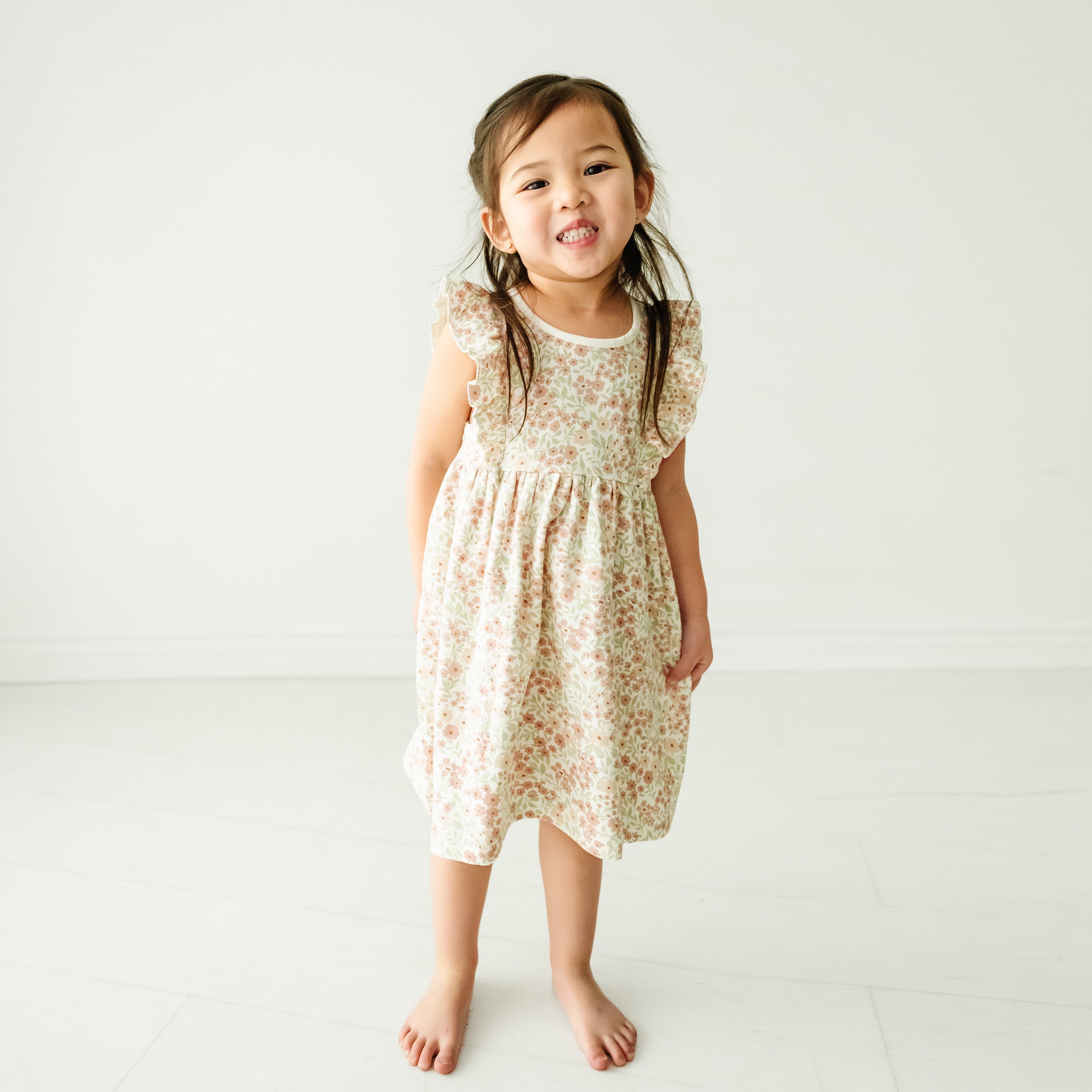 A toddler with long hair in a Organic Flutter Dress - Summer Floral by Makemake Organics stands smiling in a bright, white studio room.