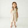 A toddler with long hair in a Organic Flutter Dress - Summer Floral by Makemake Organics stands smiling in a bright, white studio room.