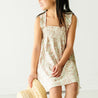 A young girl in an Organic Smocked Dress - Summer Floral by Makemake Organics, smiling to her side, holding a straw hat, standing against a white background.