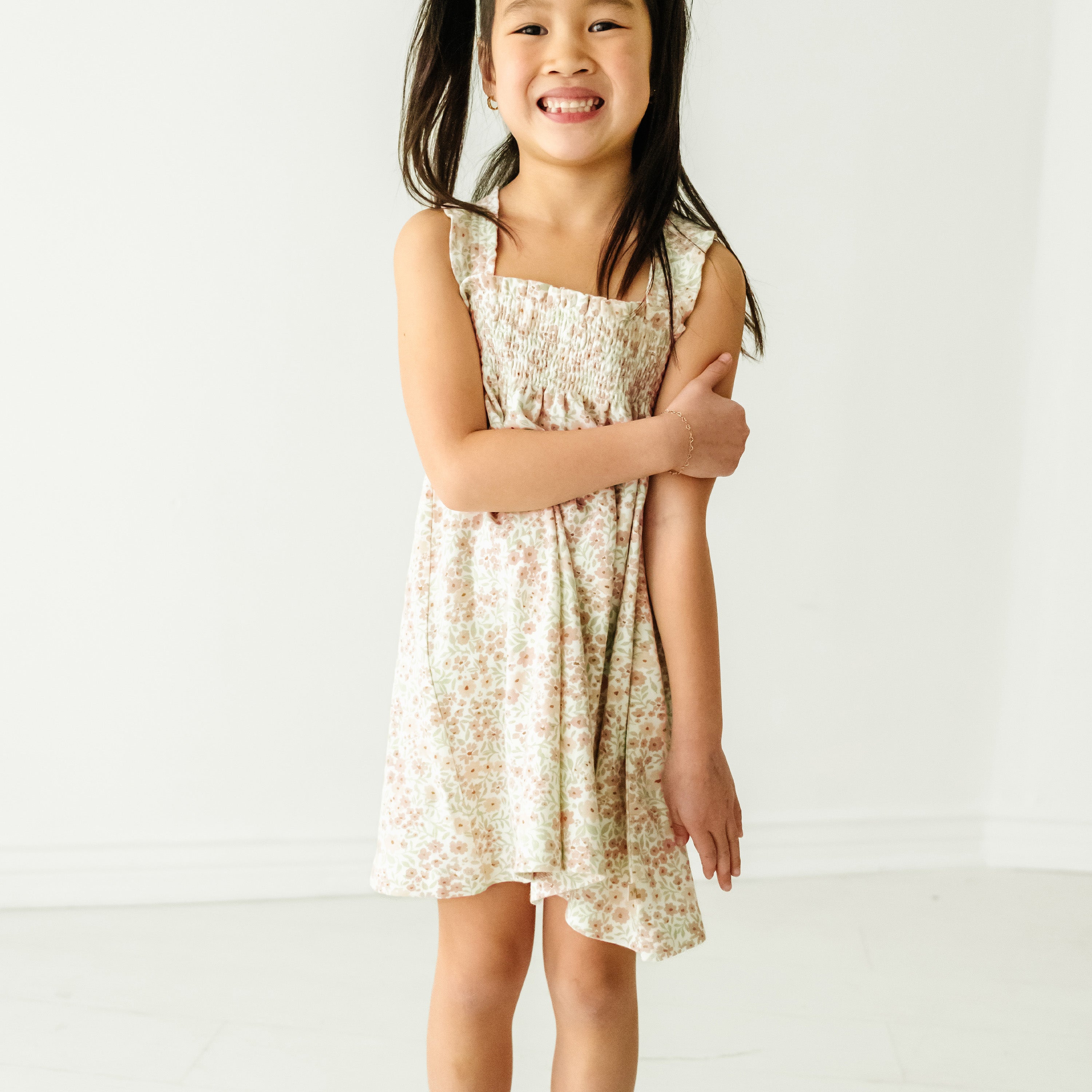 A young smiling boy with his arms crossed stands wearing a Organic Smocked Dress - Summer Floral from Makemake Organics in a bright room with a plain white background.
