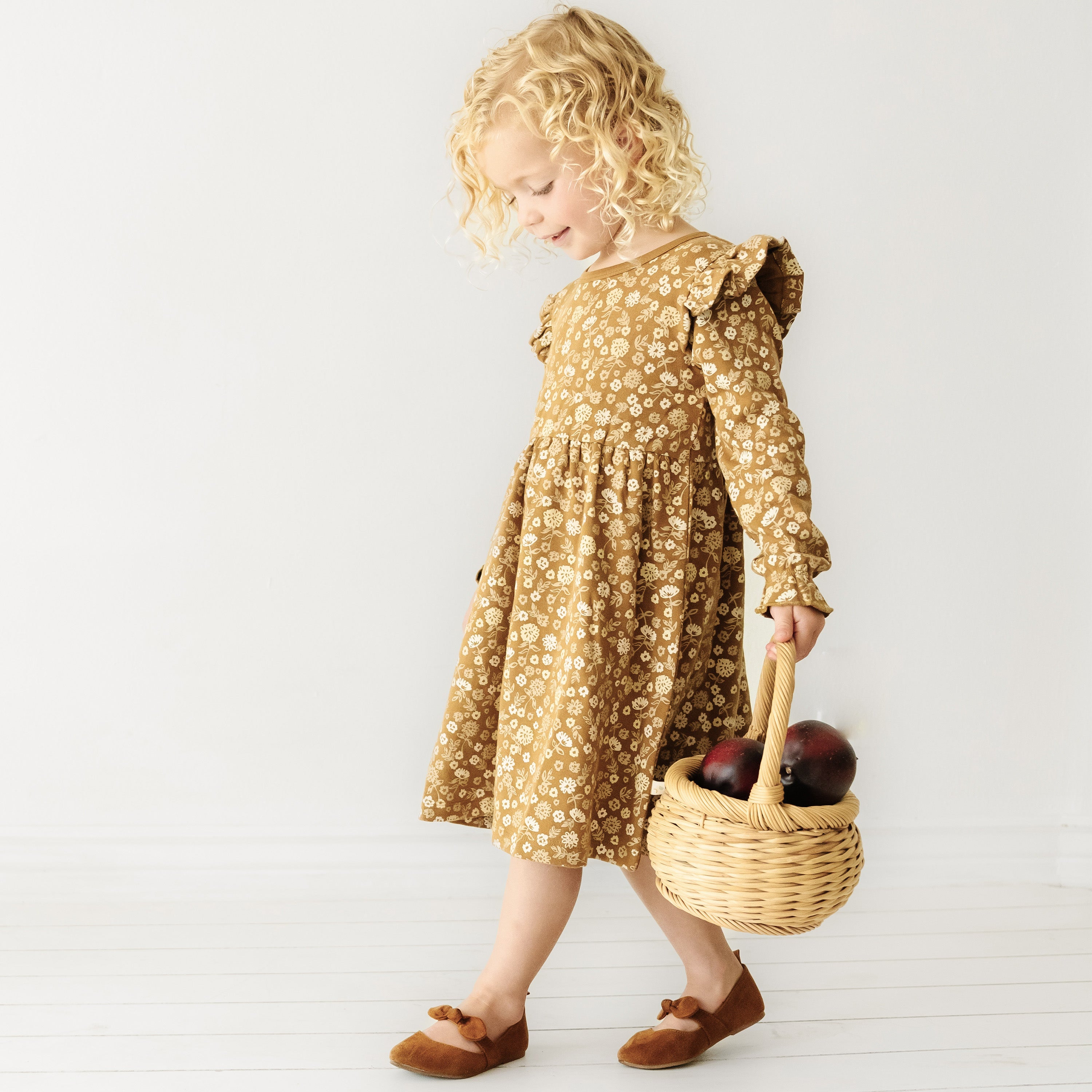 A young girl with curly blonde hair, wearing an Organic Girls Wildflower floral print dress and brown shoes, walks while holding a wicker basket filled with apples.
