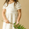A young girl in a white Organic Kids dress with gold leopard prints holds a fern leaf and a small metal hoop, with only her torso and arms visible against a beige background.