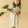 A young girl in an Organic Kids Organic Puff Sleeve Dress - Wild Safari smiling and holding a large monstera leaf, standing against a soft beige background.