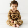 A baby with a serious expression sits on a white floor, wearing an Organic Baby Wildflower gold and brown Organic Kimono Onesie & Pants Set, holding a wooden ball.