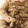 A baby in a floral Makemake Organics outfit holding a wooden mushroom toy. The close-up focuses on the baby's hands and the toy, detailing the texture of the fabric and wood.