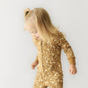A toddler with light hair, wearing the Organic 2-Way Zip Romper - Wildflower by Organic Baby, looking down as she takes a step, against a plain white background.