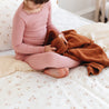 A young child in a pink pajama set sitting on a bed covered with Makemake Organics' Organic Cotton Sheet Set - Bloom, holding an orange blanket. the setting is cozy with a peaceful ambiance.