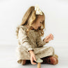 A young girl with curly hair and a bow sits cross-legged, playing with a wooden toy from Makemake Organics on a white background. She wears an Organic Ruffle Dress in Vintage Bloom from Organic Girls and brown boots, expressing her unique style.