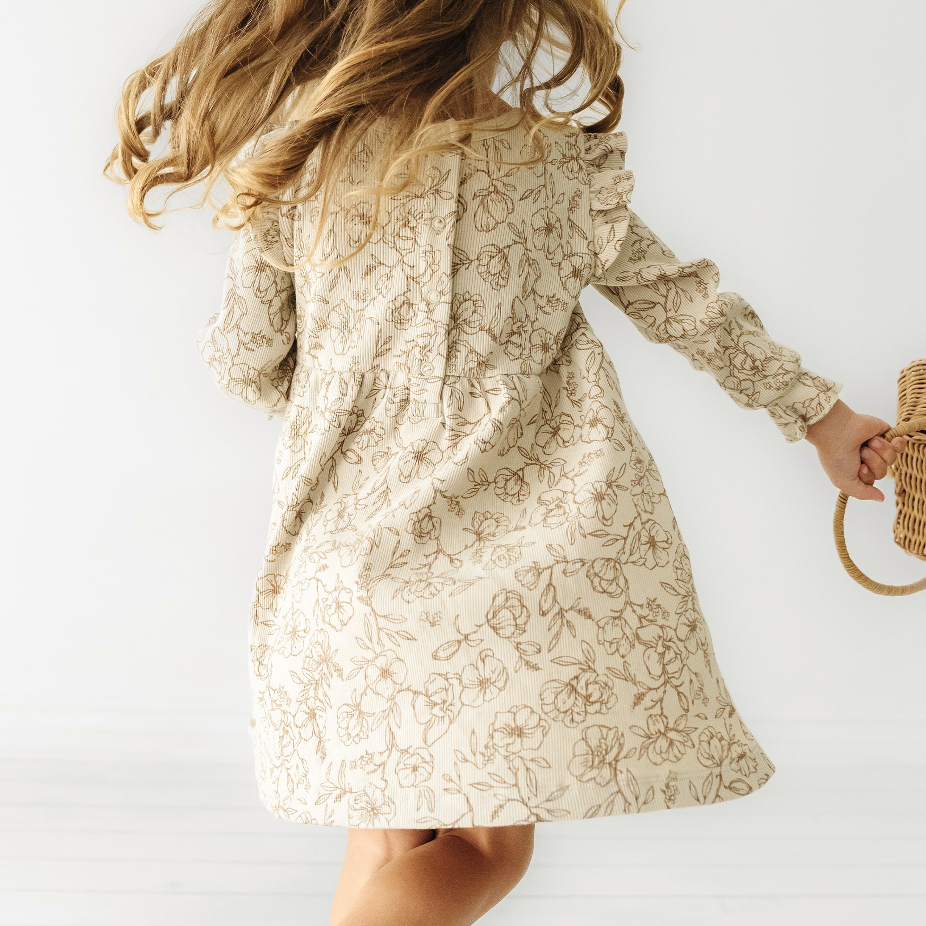 A young girl from the back wearing a Organic Girls Vintage Bloom floral dress and holding a wicker bag, her hair caught in motion, against a bright white background.