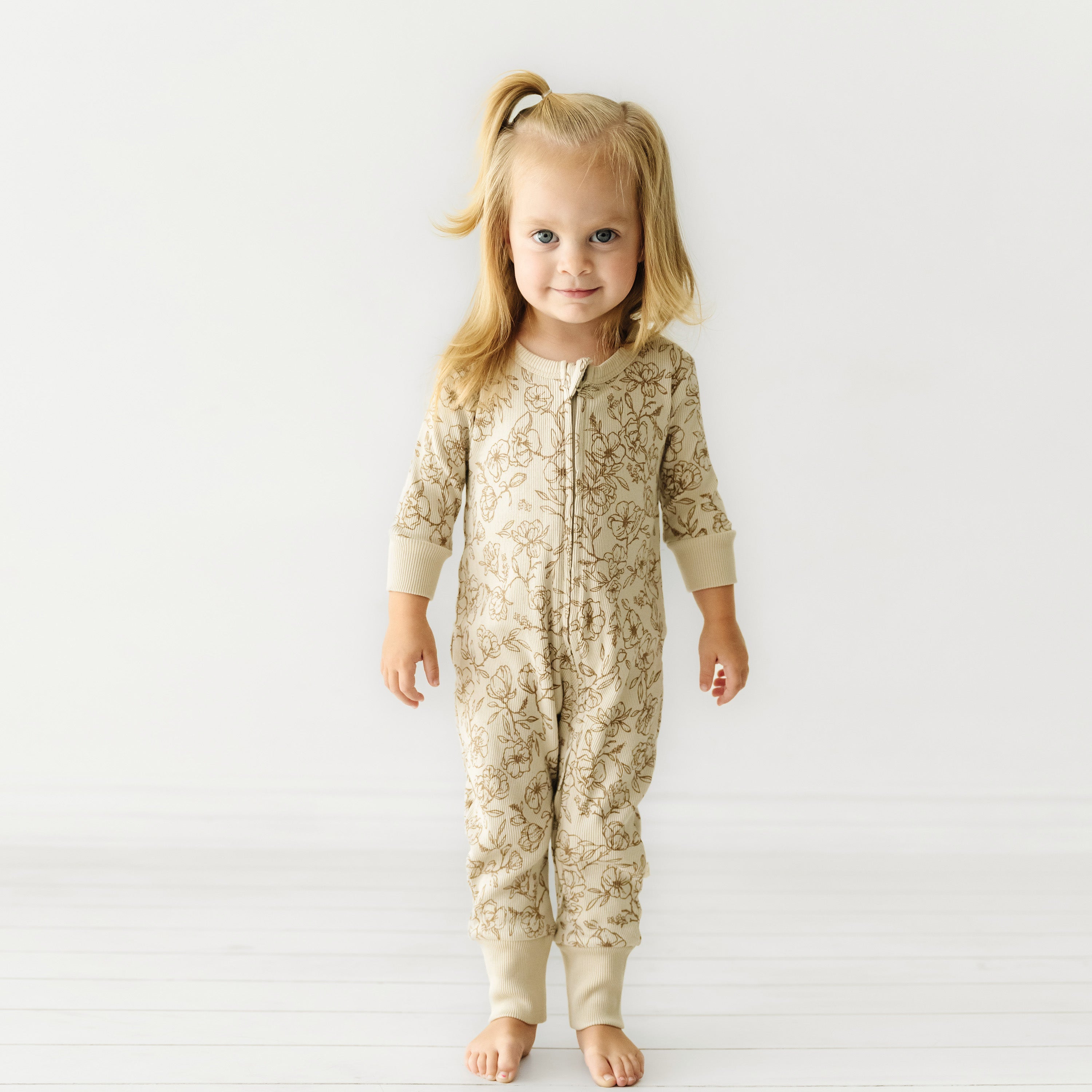 A toddler with light hair styled in a ponytail stands smiling in an Organic 2-Way Zip Romper - Vintage Bloom by Organic Baby against a white background.
