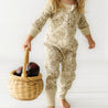 A toddler in an Organic Baby Vintage Bloom floral patterned onesie carries a wicker basket filled with large dark plums, walking barefoot on a white wooden floor.