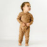 Toddler in an Organic Baby brown star-patterned Buttoned Romper - Sparkle standing barefoot on a white floor against a plain background, looking up and to the side with a curious expression.