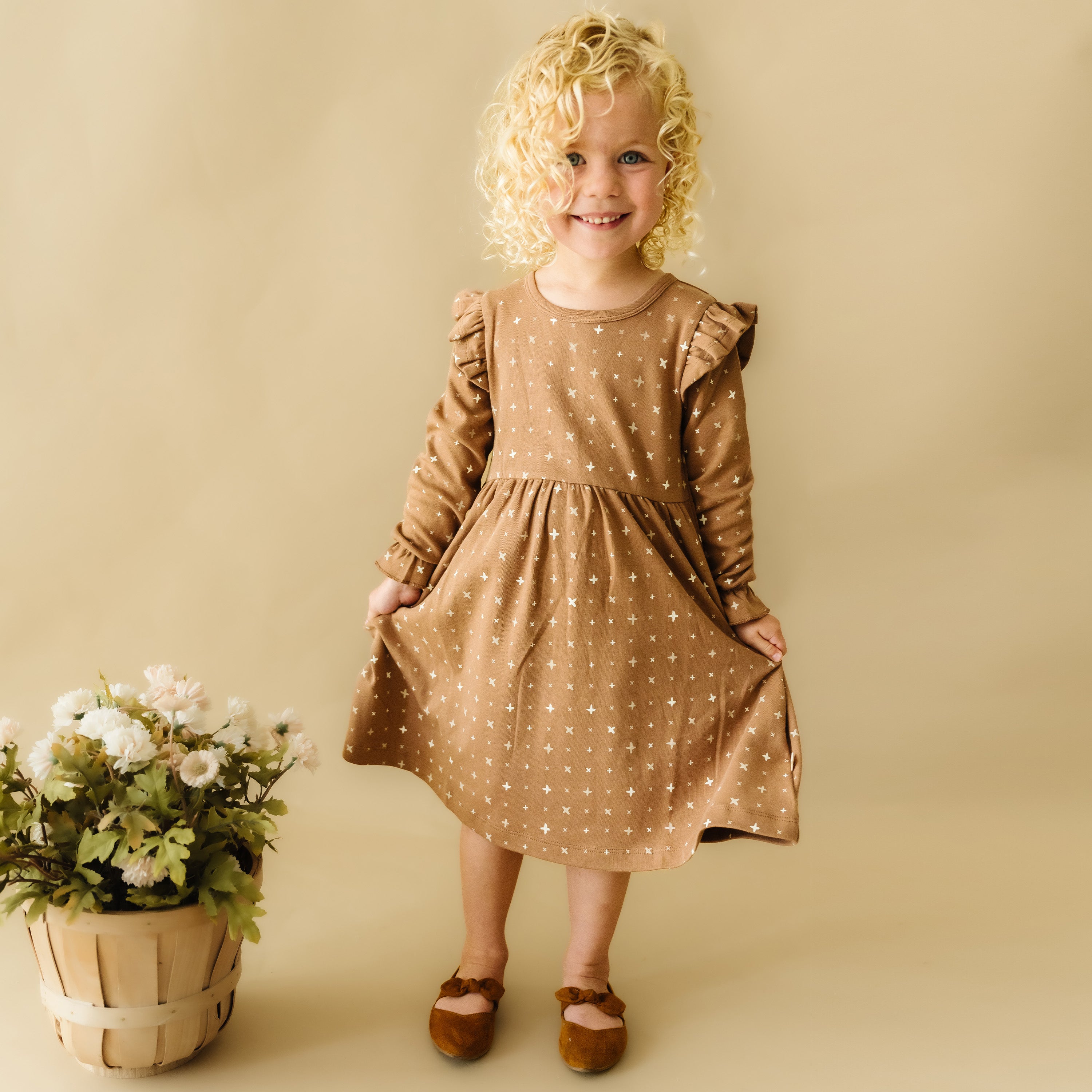 A young girl with curly blonde hair smiles while wearing an Organic Girls Sparkle Ruffle Dress and brown shoes, standing next to a basket of flowers against a tan backdrop.