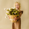 A joyful young child with curly blonde hair, wearing a brown Organic Ruffle Dress - Sparkle from Organic Girls, holds a large wooden basket filled with white and pink flowers against a beige background.