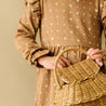 A close-up image of a person holding a Makemake Organics Organic Ruffle Dress - Sparkle wicker basket, wearing a brown dress with white star patterns, against a beige background. Only the midsection and hands are visible