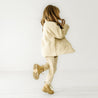 A young girl in an Organic Kids cream quilted jacket and pants from Makemake Organics walks away from the camera. Her long brown hair is visible as she walks on her toes, showcasing her tan boots.