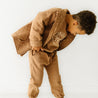 A young child dressed in a cozy Organic Kids brown outfit playfully bends forward while looking down, set against a plain white background.