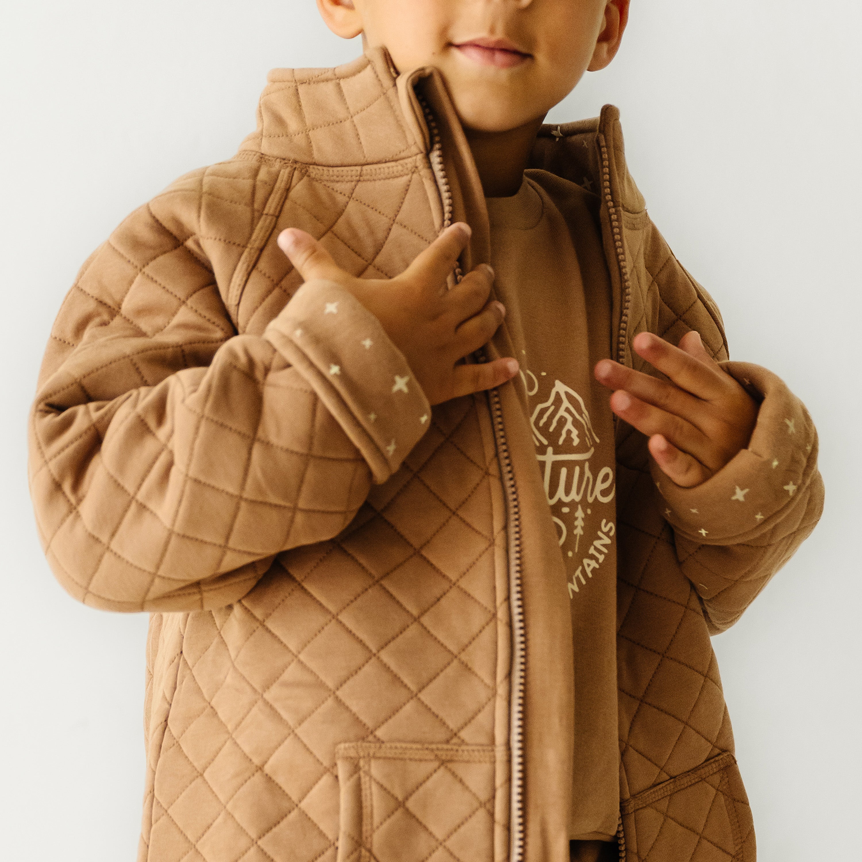 A young child in a brown Organic Kids Organic Merino Wool Zip Jacket - Cocoa over a hoodie gives a thumbs-up gesture. Only the lower half of the child's face is visible. The background is plain and