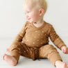 A toddler in an Organic Baby brown Kimono Onesie & Pants Set - Sparkle adorned with white stars from Makemake Organics sits on the floor, looking to the side with a curious expression. The background is plain and light-colored.