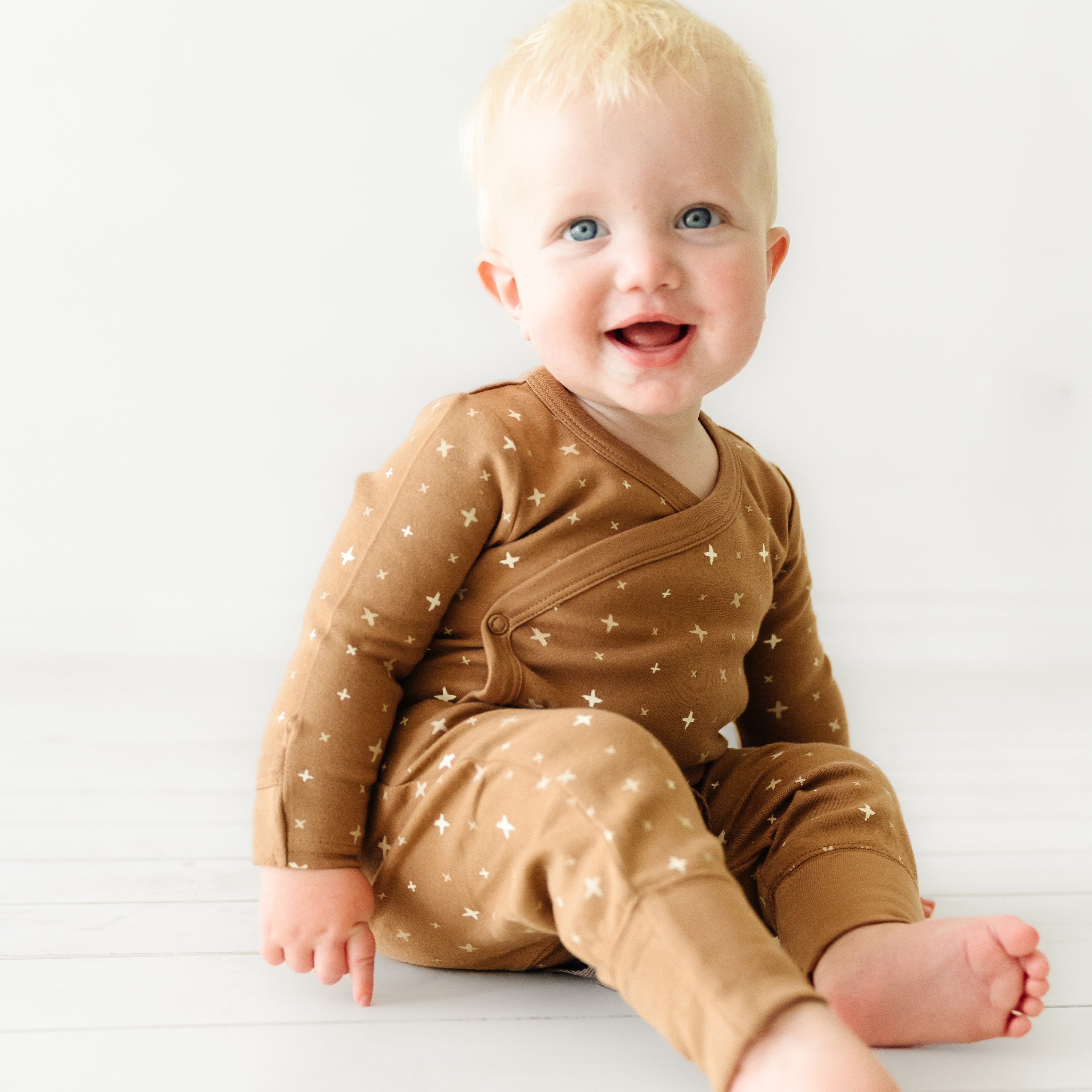 A joyful blonde baby with blue eyes wearing an Organic Baby brown kimono onesie and pants set decorated with stars, sitting on a white floor and smiling.