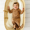 A joyful baby wearing an Organic Baby star-patterned brown kimono knotted sleep gown lies in a woven basket, smiling widely against a white wooden floor background.