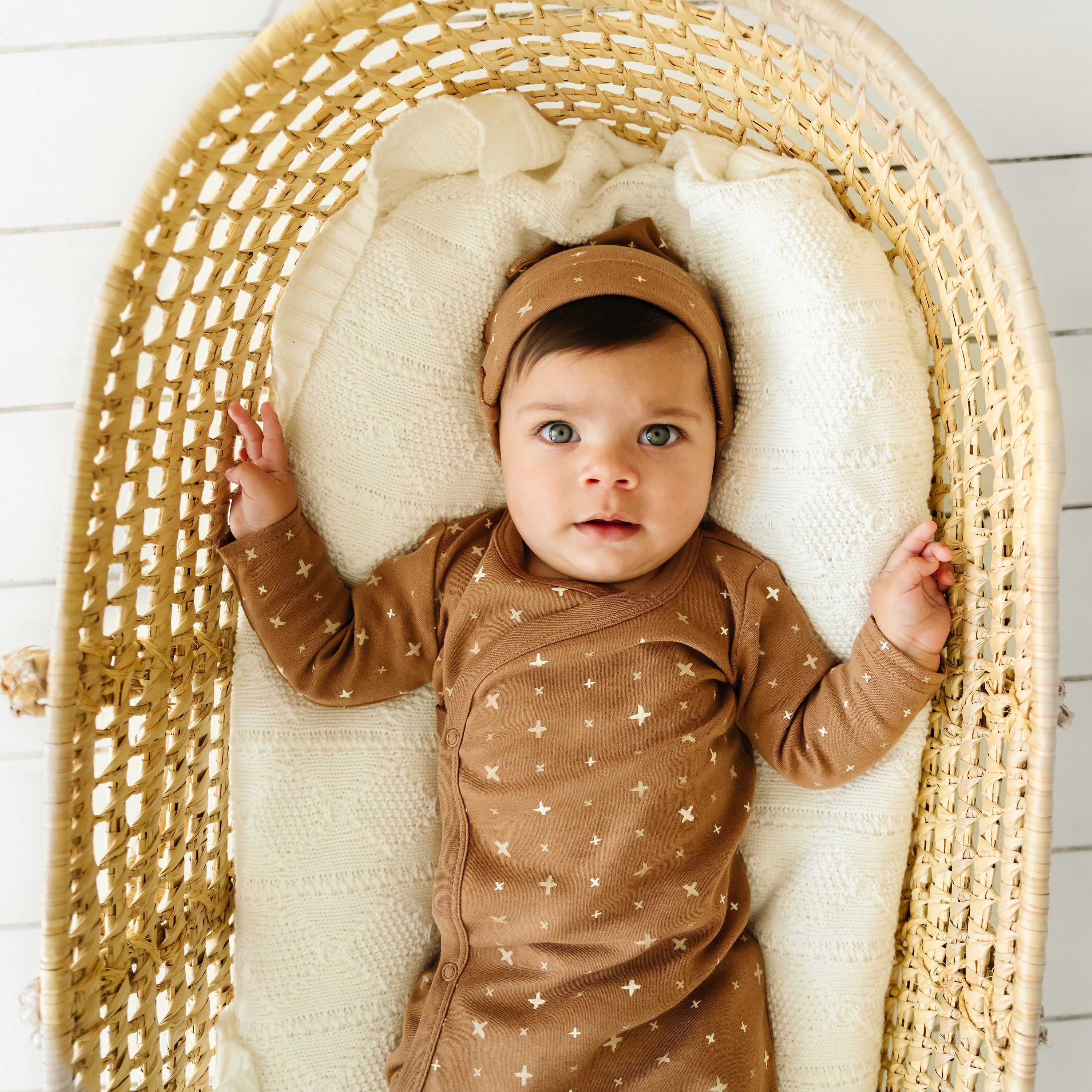 A baby with wide eyes lies in a woven bassinet, wearing a beige onesie from Makemake Organics with gold stars, under a cream-colored blanket. The baby is also wearing a matching Organic Baby Organic Kimono Knotted Sleep Gown - Sparkle.