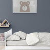 A cozy children's bedroom with a white bed covered in Makemake Organics' Organic Cotton Sheet Set - Cobi Blue Stripes, surrounded by pillows. a framed picture of a cartoon bear hangs on the gray wall, and a wooden toy house sits nearby.