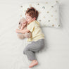 A young boy asleep in a comfortable position on a white bed, holding a teddy bear and resting his head on an Organic Cotton Toddler Pillowcase - Rainbow from Makemake Organics. he is wearing a yellow shirt and gray pants.