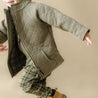 A toddler in motion, wearing an Organic Merino Wool Zip Jacket in Olive by Organic Kids over a beige outfit and tan boots, with a playful and joyful expression, set against a matching green background.