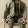 A young child wearing an Organic Kids Organic Merino Wool Zip Jacket in Olive and green plaid pants, sitting on a wooden stool with a neutral background. Only the lower half of the face and body are visible.