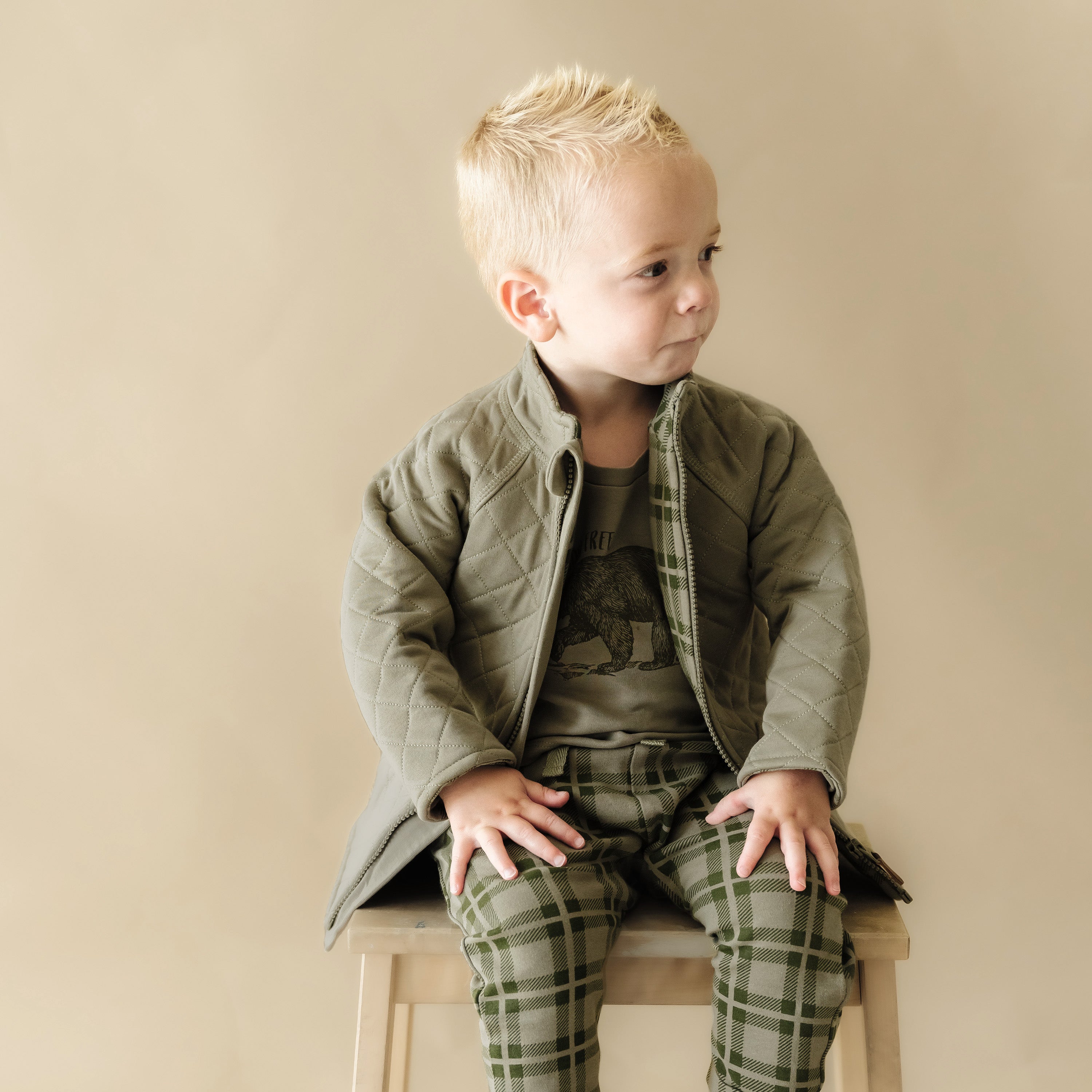 A young boy with blond hair sits on a wooden stool, stylishly dressed in Organic Kids' Organic Merino Wool Zip Jacket in Olive, camo shirt, and plaid pants, looking off to the side thoughtfully.