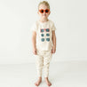 A baby wearing oversized orange sunglasses and a beige t-shirt with sunglass prints stands confidently in a white studio.