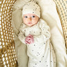 A toddler wearing a Makemake Organics Organic Kimono Knotted Sleep Gown in Pixie Dots and matching hat lies in a woven bassinet, looking upward with a curious expression.