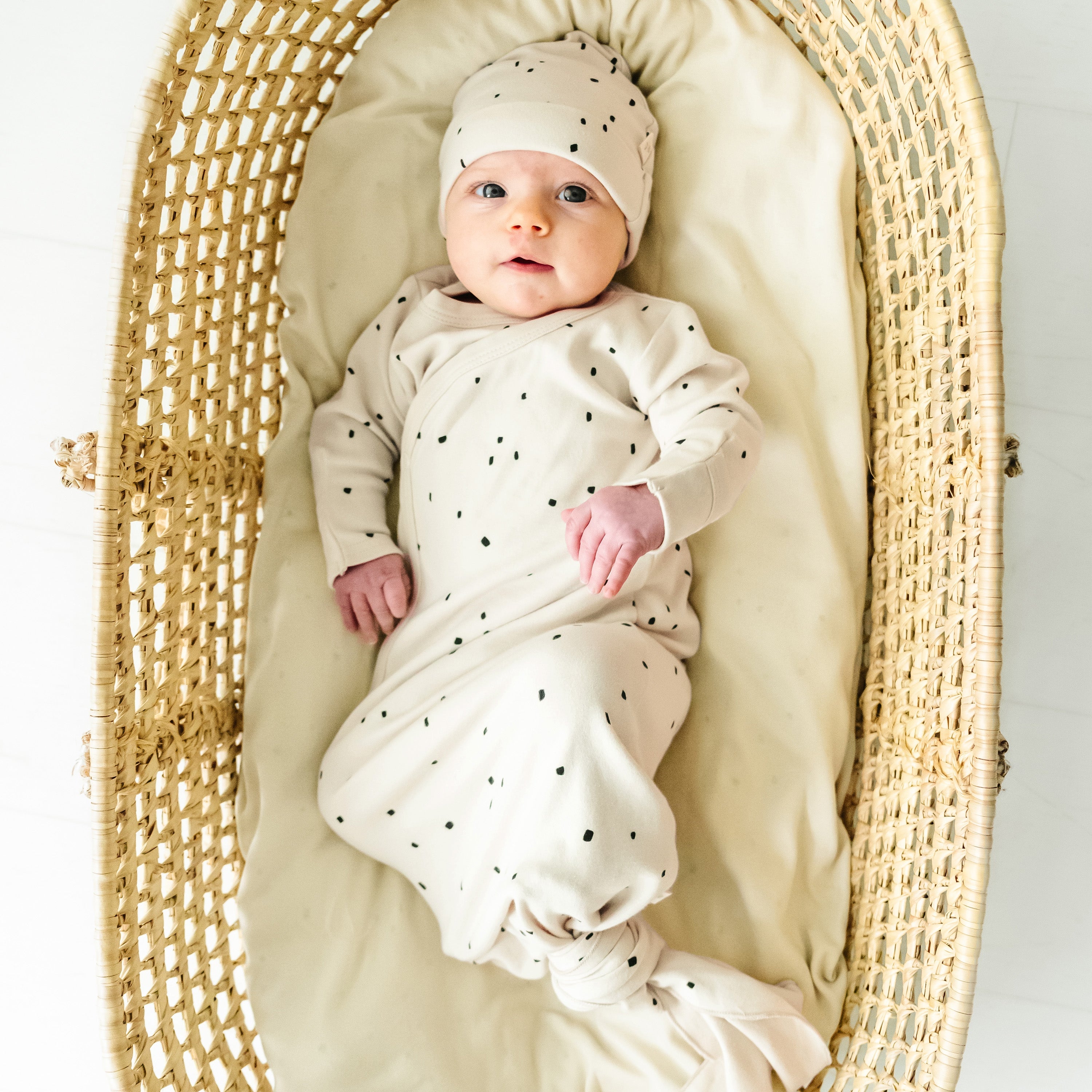 A toddler wearing a Makemake Organics Organic Kimono Knotted Sleep Gown in Pixie Dots and hat lies in a woven, oval-shaped bassinet, looking upward with a serene expression on a white background.