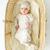 A toddler wearing a Makemake Organics Organic Kimono Knotted Sleep Gown in Pixie Dots and hat lies in a woven, oval-shaped bassinet, looking upward with a serene expression on a white background.