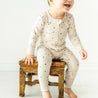 A joyful baby in a Makemake Organics Organic 2-Way Zip Romper - Pixie Dots sits on a rustic wooden stool, laughing in a bright, airy room.