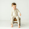 A baby with a side-swept hairstyle is sitting on a small vintage wooden stool, wearing an Organic 2-Way Zip Romper - Pixie Dots from Makemake Organics, pointing downwards.