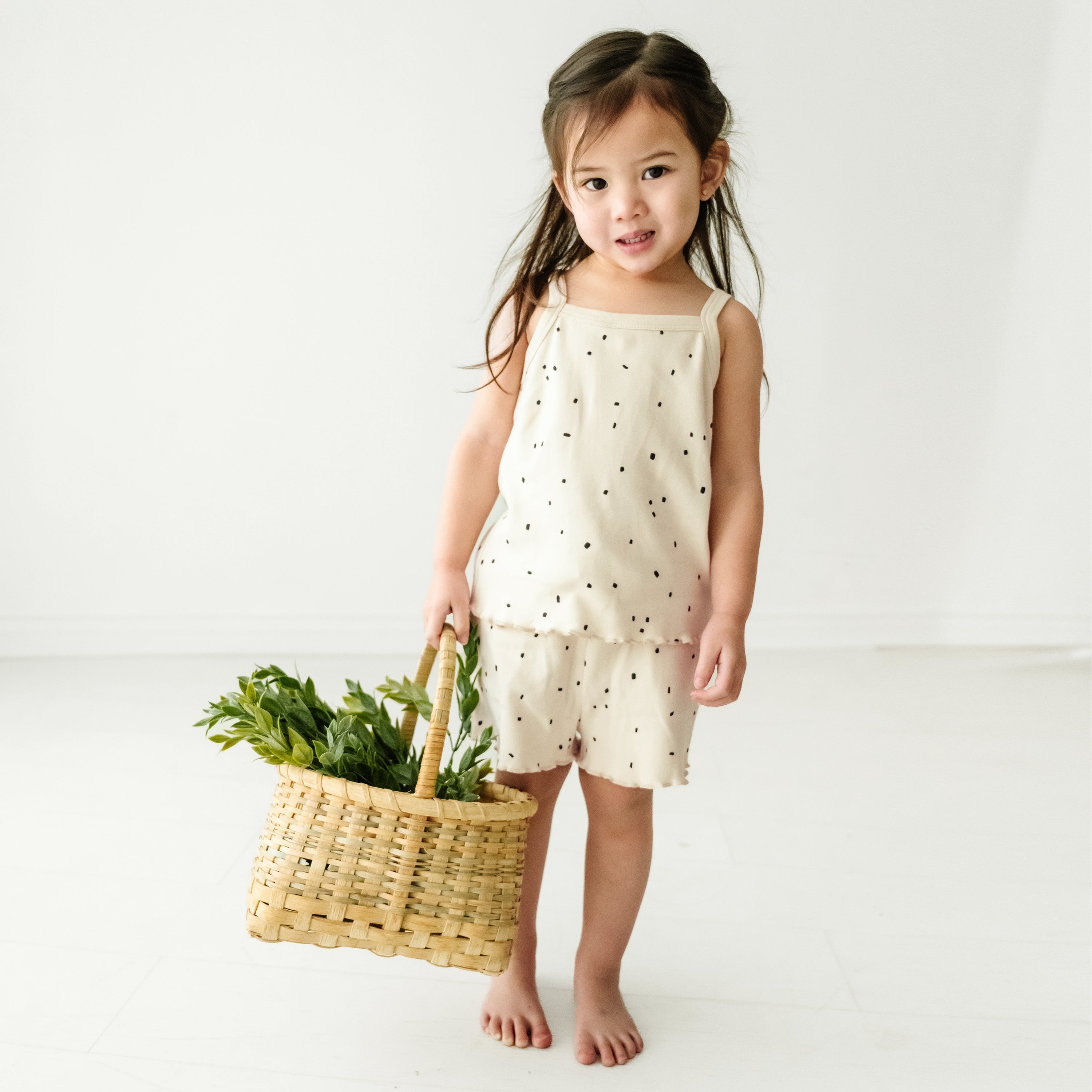 A toddler in a Organic Spaghetti Top & Shorts Set - Pixie Dots by Makemake Organics holds a woven basket filled with fresh green vegetables, standing against a white background, looking happy.