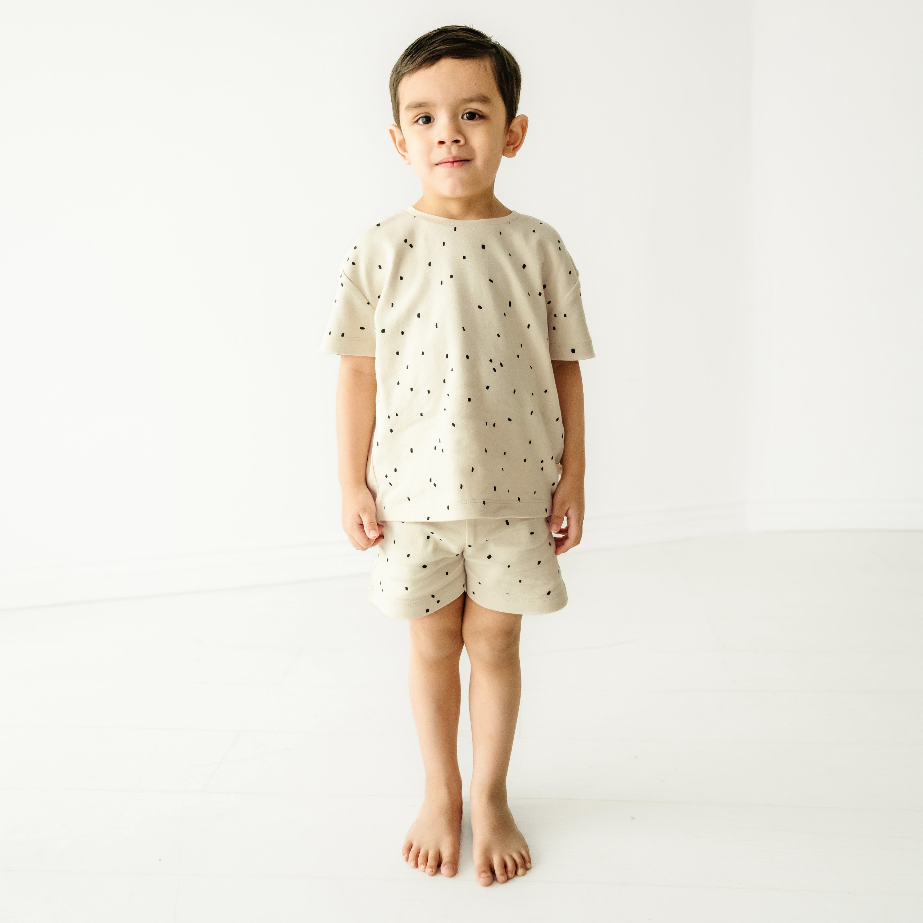 A young child wearing a Pixie Dots Organic Tee and Shorts Set from Makemake Organics stands barefoot in a bright, plain white room, looking directly at the camera.