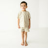 A young child wearing a Pixie Dots Organic Tee and Shorts Set from Makemake Organics stands barefoot in a bright, plain white room, looking directly at the camera.