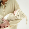 A woman cradling a sleeping baby wrapped in a Makemake Organics Pixie Dots swaddle blanket and hat, with a focus on the tender embrace of their arms against a soft, neutral background.