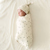 A newborn baby sleeps peacefully, swaddled in a white Makemake Organics organic swaddle blanket adorned with black Pixie Dots, wearing a matching hat, on a soft, light-colored bedsheet.